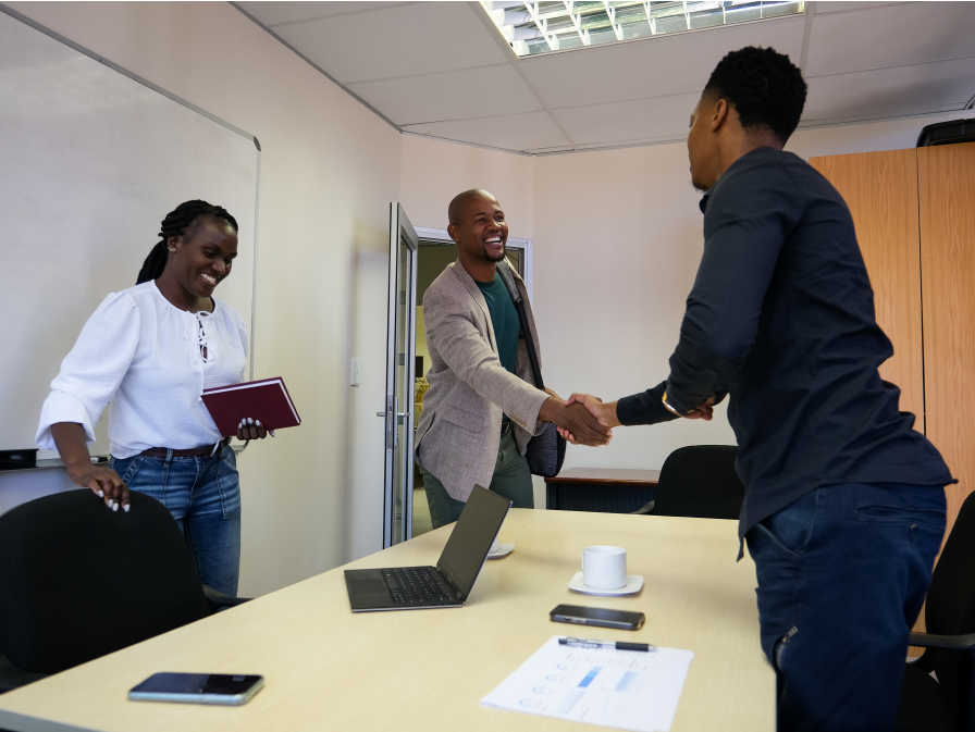 Three colleagues in a conference room. Two men are shaking hands and smiling, while a woman stands nearby holding a notebook and smiling. A laptop and documents are on the table.