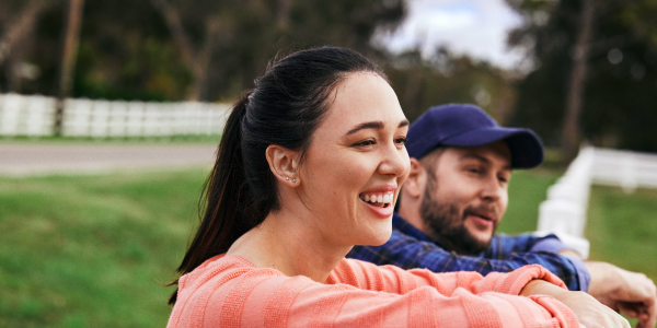 Smiling woman in a pink sweater and man in a blue hat leaning on a white fence, with greenery and trees in the background.