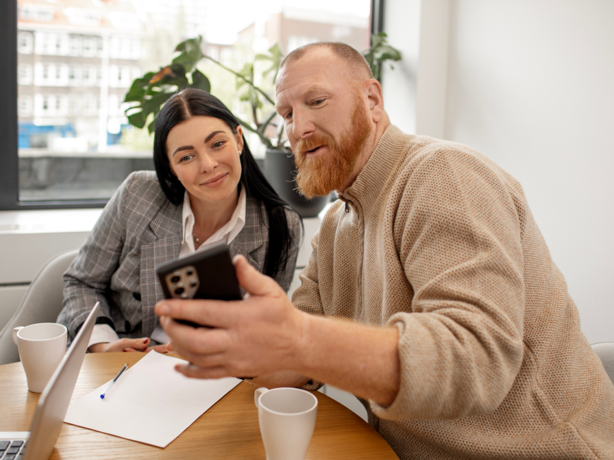 Two colleagues are in an office sitting at a table, looking at a smartphone. The woman is wearing a gray blazer, and the man has a beard and is wearing a beige sweater.