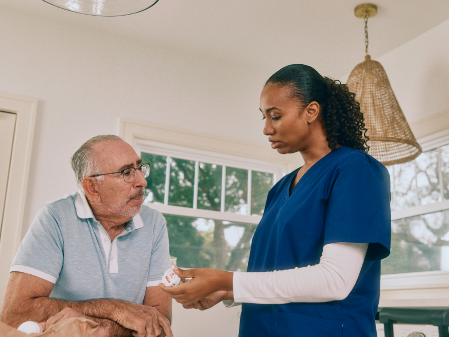 A nurse in blue scrubs is talking to an older man in a light blue polo shirt, holding a prescription bottle.