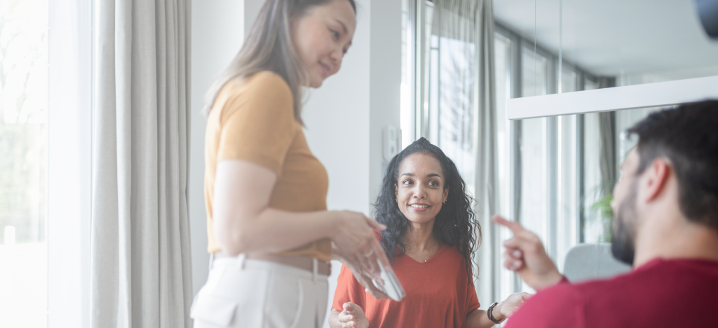 Three colleagues in a modern office: a woman with a tablet, a smiling woman listening, and a man gesturing as he speaks.