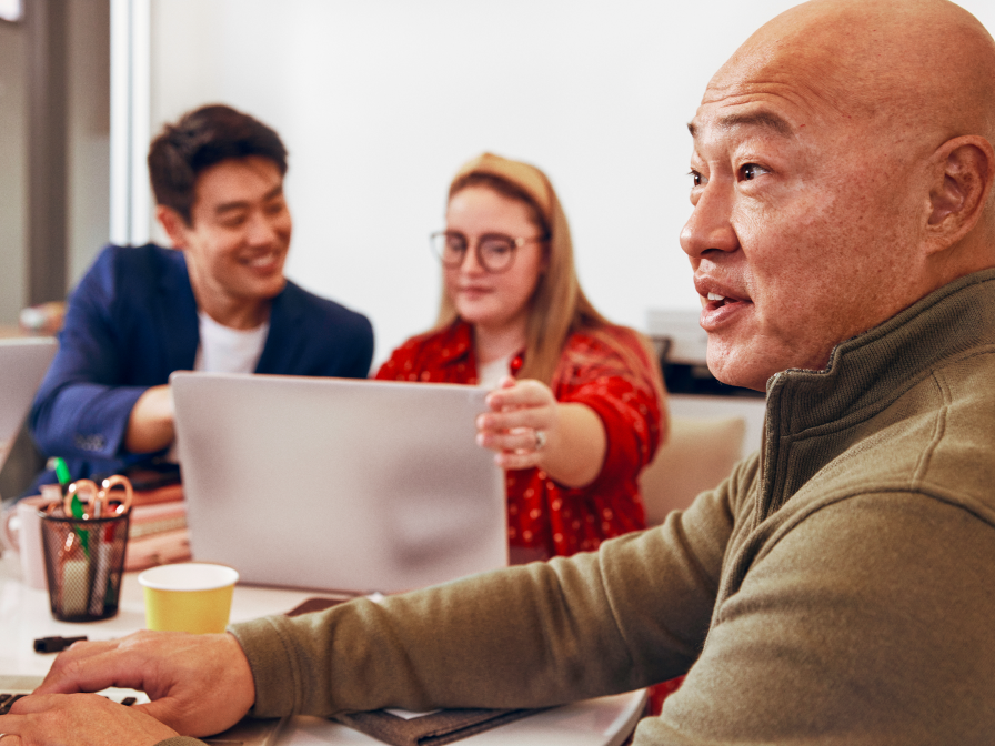 Three colleagues working together in an office. An older man in a green sweater is speaking, while a younger man in a navy suit and a woman in a red cardigan are looking at a laptop screen, smiling.